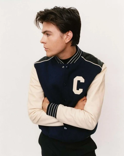 Portrait of a young adult male wearing a navy and cream varsity jacket, arms crossed, and looking off-camera against a white backdrop.