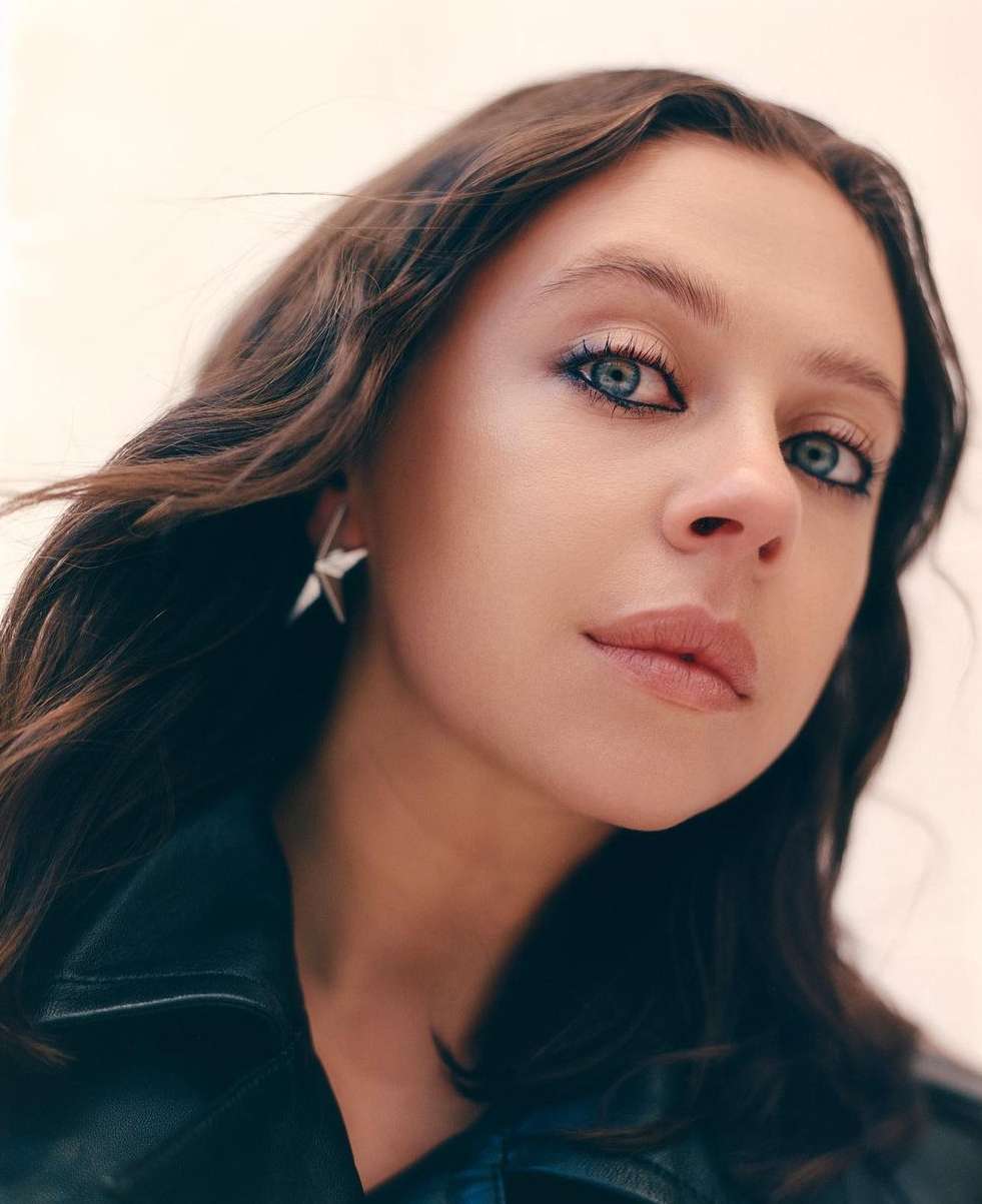 Close-up portrait of a young adult woman with wavy brown hair, blue eye makeup, a black jacket, and star-shaped earrings.