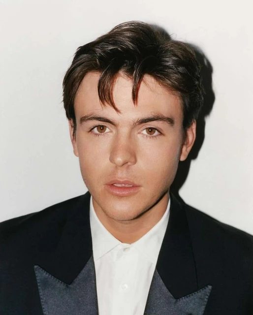 Portrait of a young adult male with brown hair, wearing a black tuxedo jacket and white shirt against a plain white background.