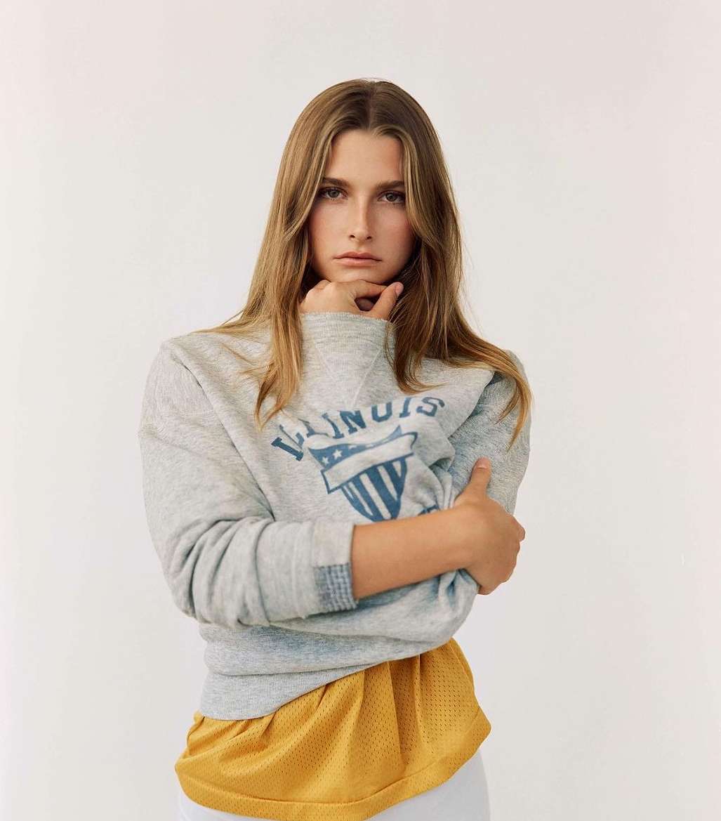 Blonde woman wearing grey 'Illinois' sweatshirt poses in a brightly lit studio with arms crossed, looking directly at the camera.
