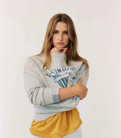 Blonde woman wearing grey 'Illinois' sweatshirt poses in a brightly lit studio with arms crossed, looking directly at the camera.