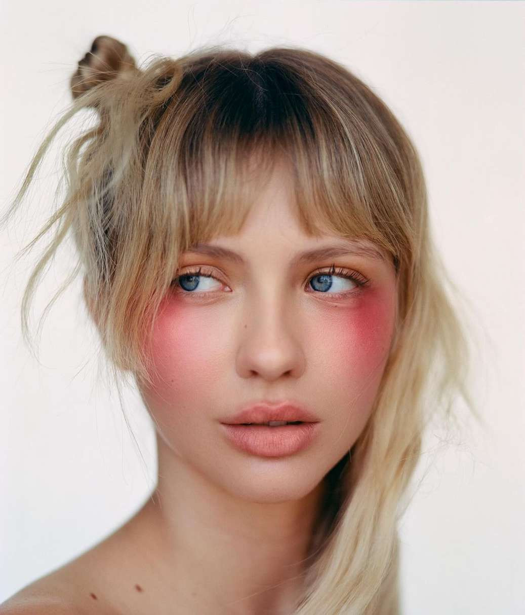 Close-up portrait of a woman with blonde hair, bangs, and bright pink blush across her cheeks against a light background.