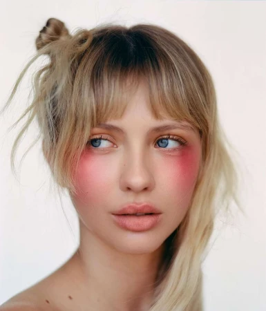 Close-up portrait of a woman with blonde hair, bangs, and bright pink blush across her cheeks against a light background.