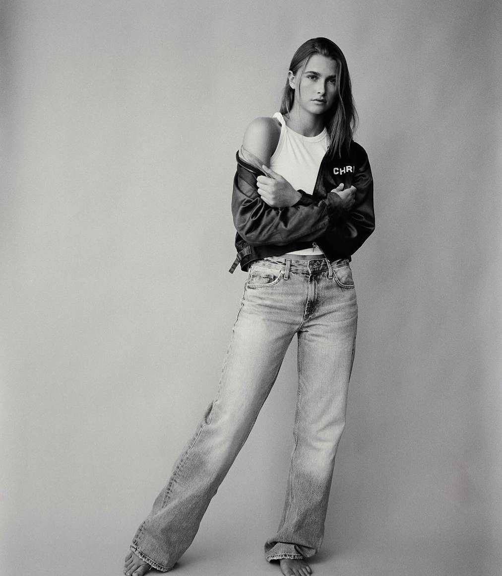 Full body shot of a woman in jeans, tank top, and jacket, standing indoors with bare feet, in a monochrome studio photograph.