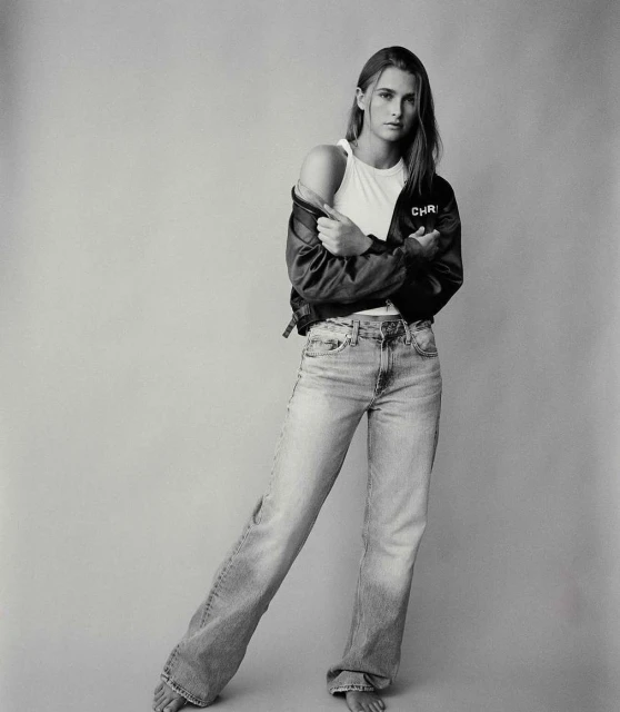 Full body shot of a woman in jeans, tank top, and jacket, standing indoors with bare feet, in a monochrome studio photograph.
