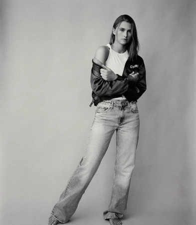 Full body shot of a woman in jeans, tank top, and jacket, standing indoors with bare feet, in a monochrome studio photograph.
