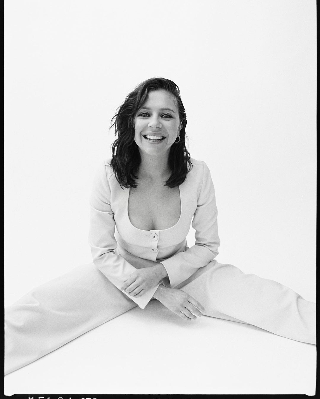 A smiling woman with dark hair sits in a cream-colored outfit against a white background, looking directly at the camera in a medium shot.