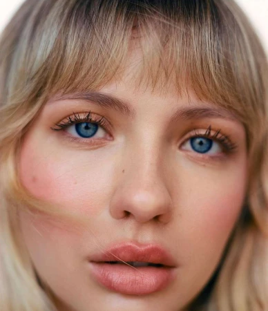 Close-up portrait of a blonde woman with blue eyes, bangs, and natural makeup against a plain background