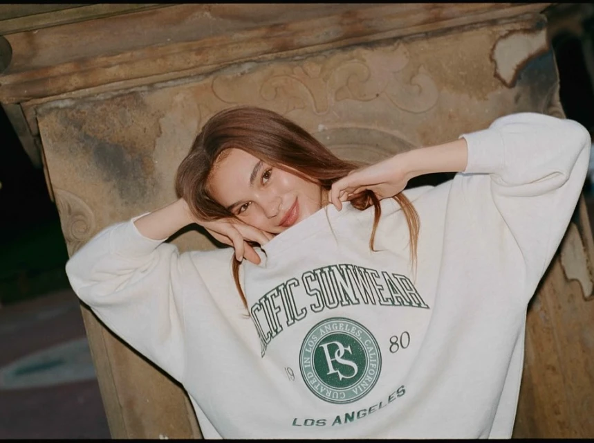 A young woman poses in front of a stone wall, wearing a vintage-style sweatshirt. She has long brown hair, a warm smile, and fair skin.