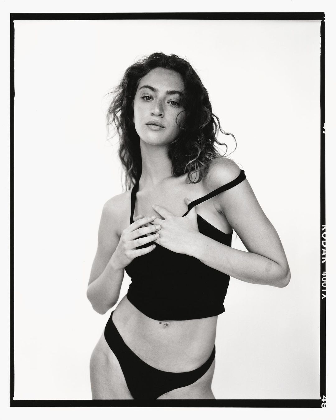 Monochrome portrait of a woman in a dark top and underwear, posed in front of a bright background, framed by a black border.