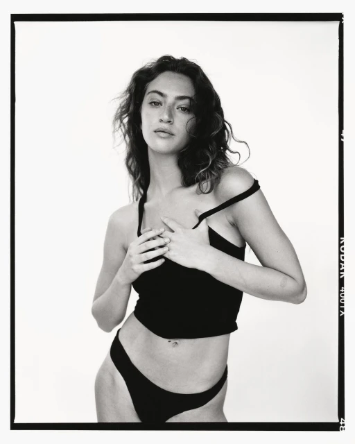 Monochrome portrait of a woman in a dark top and underwear, posed in front of a bright background, framed by a black border.
