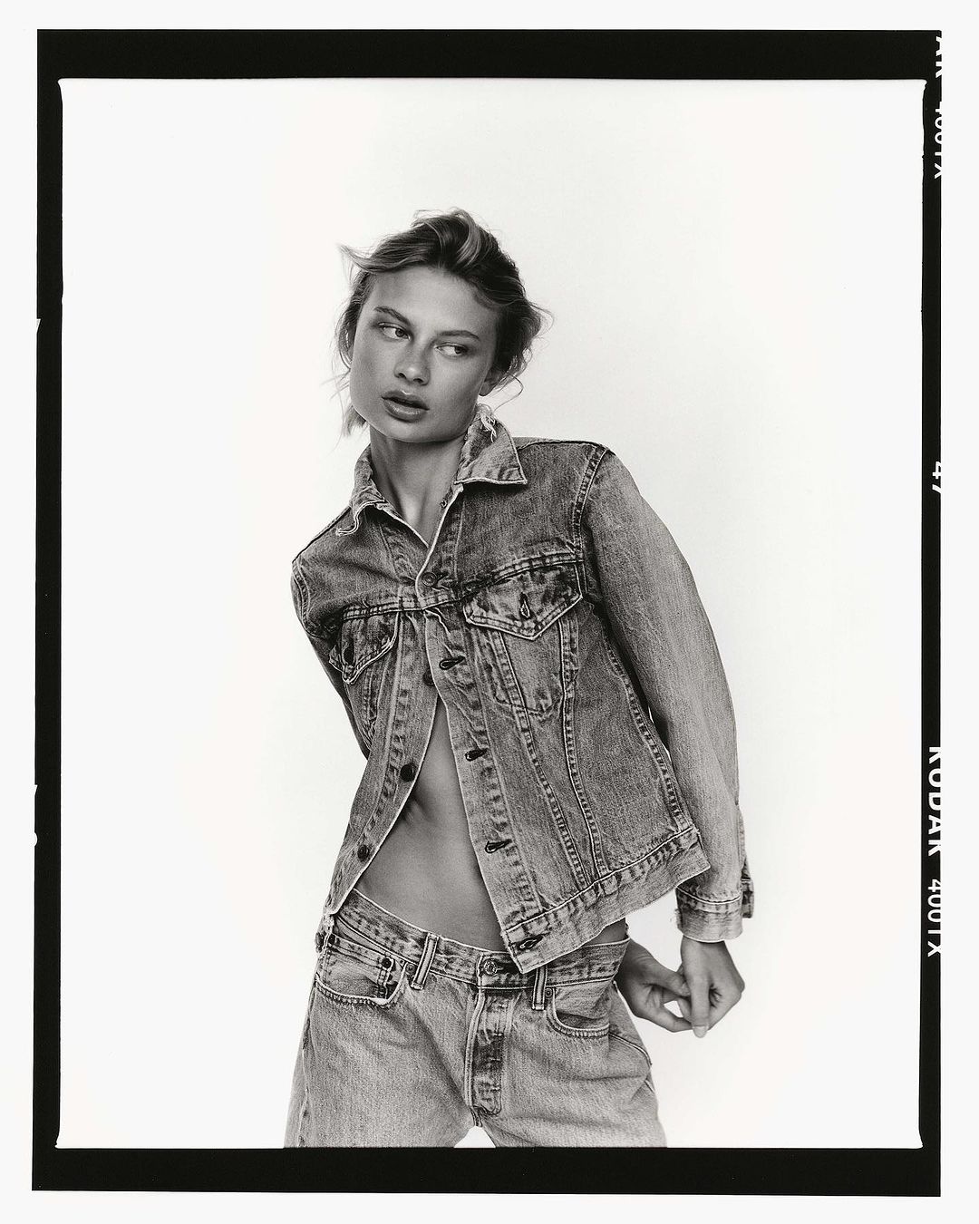 Black and white portrait of a woman wearing a denim jacket and jeans against a white backdrop.