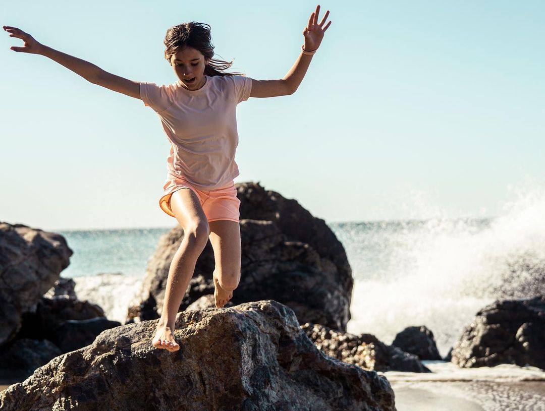 Girl jumping on rocks at the beach, ocean waves and blue sky in background, youth fashion, fitness activity, lifestyle, daylight.