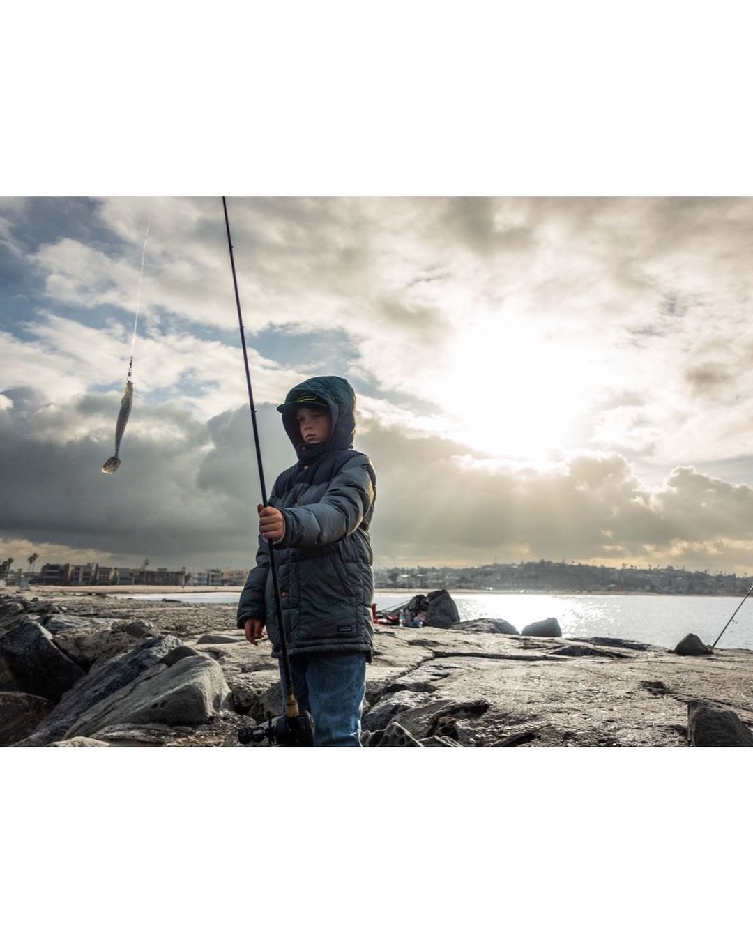 Young boy fishing off a rocky pier, holding a fishing rod with a lure, against a cloudy sky and ocean backdrop.