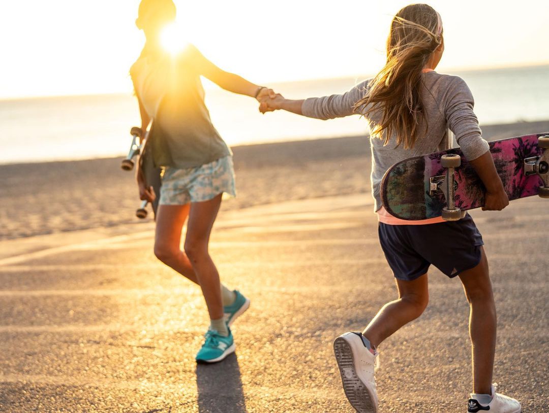 Two young girls holding hands while carrying skateboards on a paved surface, bathed in golden sunlight near a body of water.