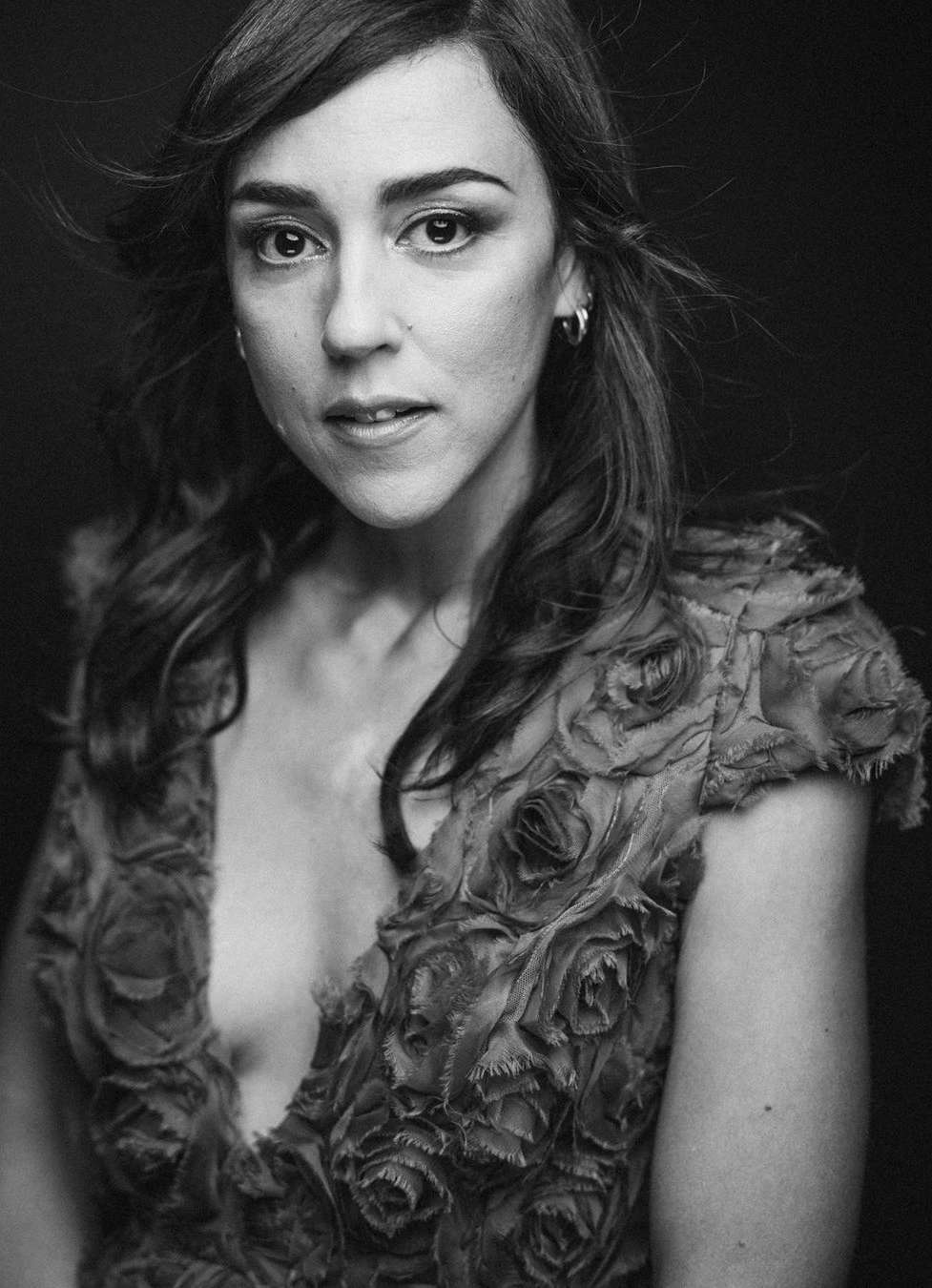 Black and white portrait of a woman with shoulder-length brunette hair, wearing a top adorned with rose-like embellishments, against a dark background.