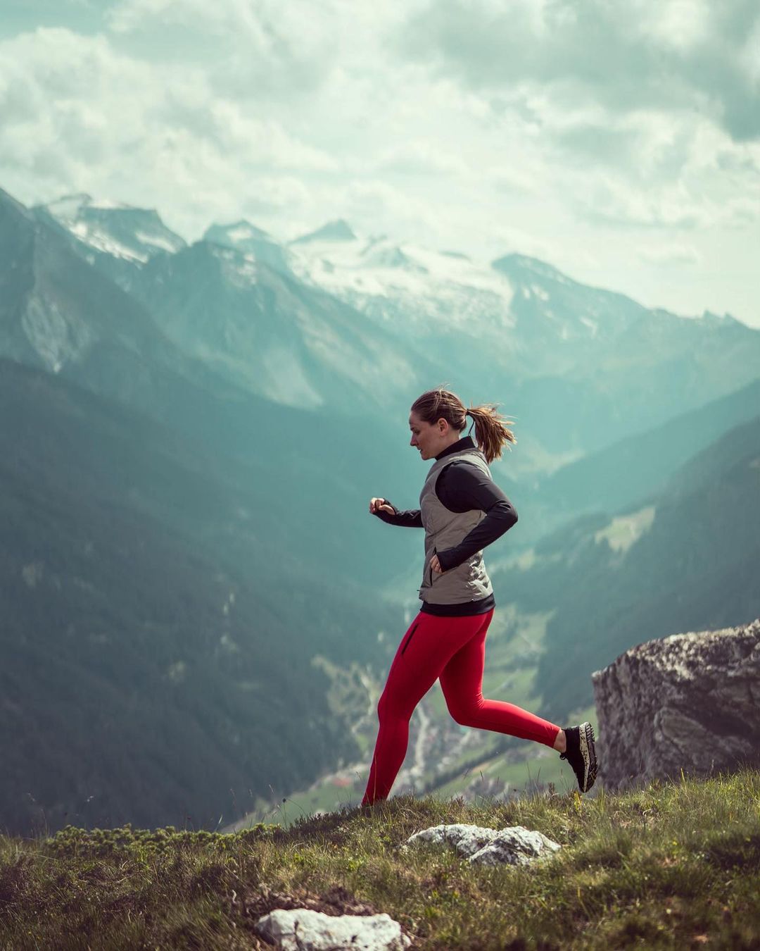 Adult woman trail running outdoors in a mountainous landscape with evergreen trees and snow-capped mountains under a cloudy sky.