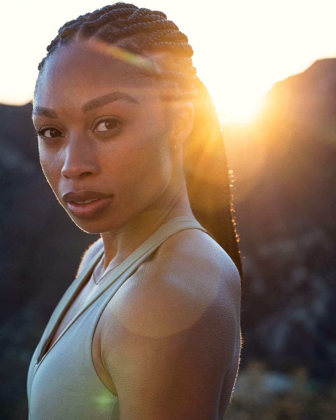 Close-up portrait of an adult woman with braided hair and a sage-green tank top, set against a sunlit mountain backdrop at sunset.