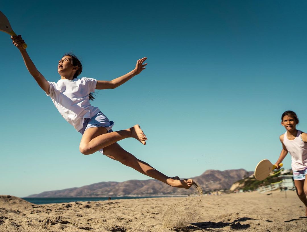 A girl jumps on a sandy beach with paddle in hand, another girl in the background and mountains across ocean under clear blue sky.