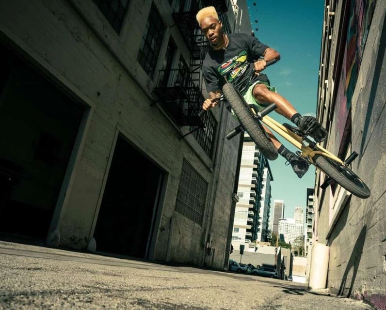 Young adult male performing a bicycle stunt in an urban alleyway, riding up a wall with city buildings in the background, action shot.
