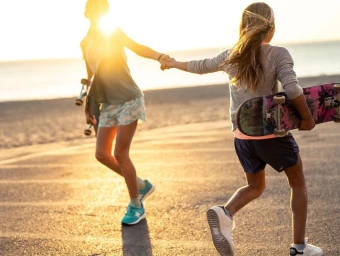 Two young people holding hands walking along a paved coastal path at sunset while carrying skateboards.
