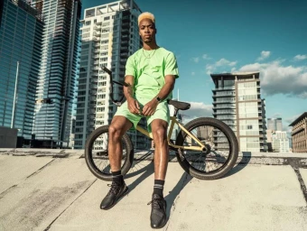 A Black man with bleached blonde hair sitting on a BMX bicycle on a city rooftop against a backdrop of high-rise buildings.