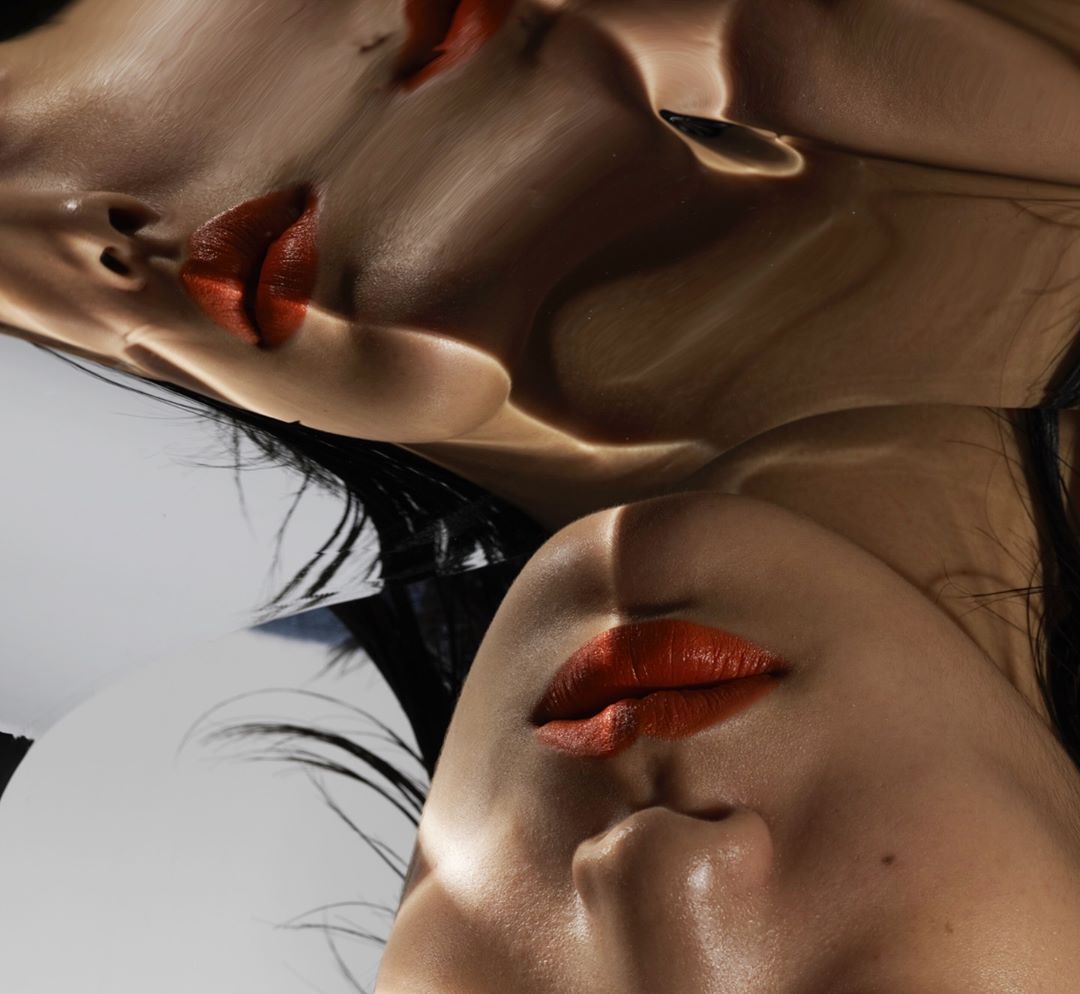 Dramatic beauty shot featuring a woman's face with red lipstick, reflected in a high-shine surface, emphasizing light and shadow.