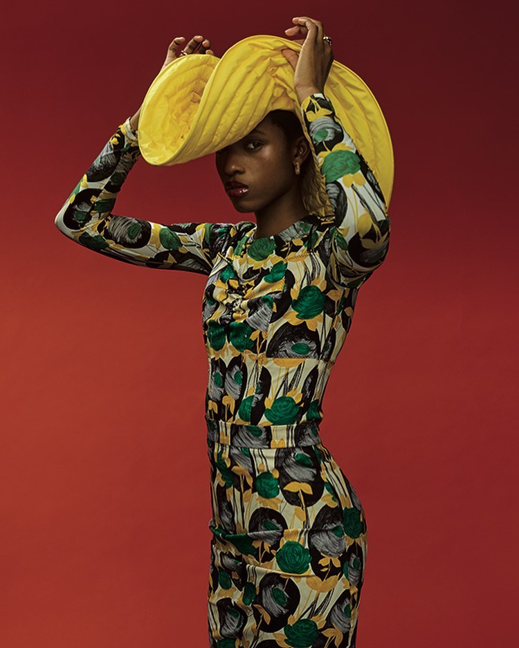 A stylish African American woman in a floral dress and yellow hat strikes a pose against a vibrant red backdrop in a fashion editorial.