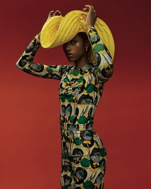 A stylish African American woman in a floral dress and yellow hat strikes a pose against a vibrant red backdrop in a fashion editorial.