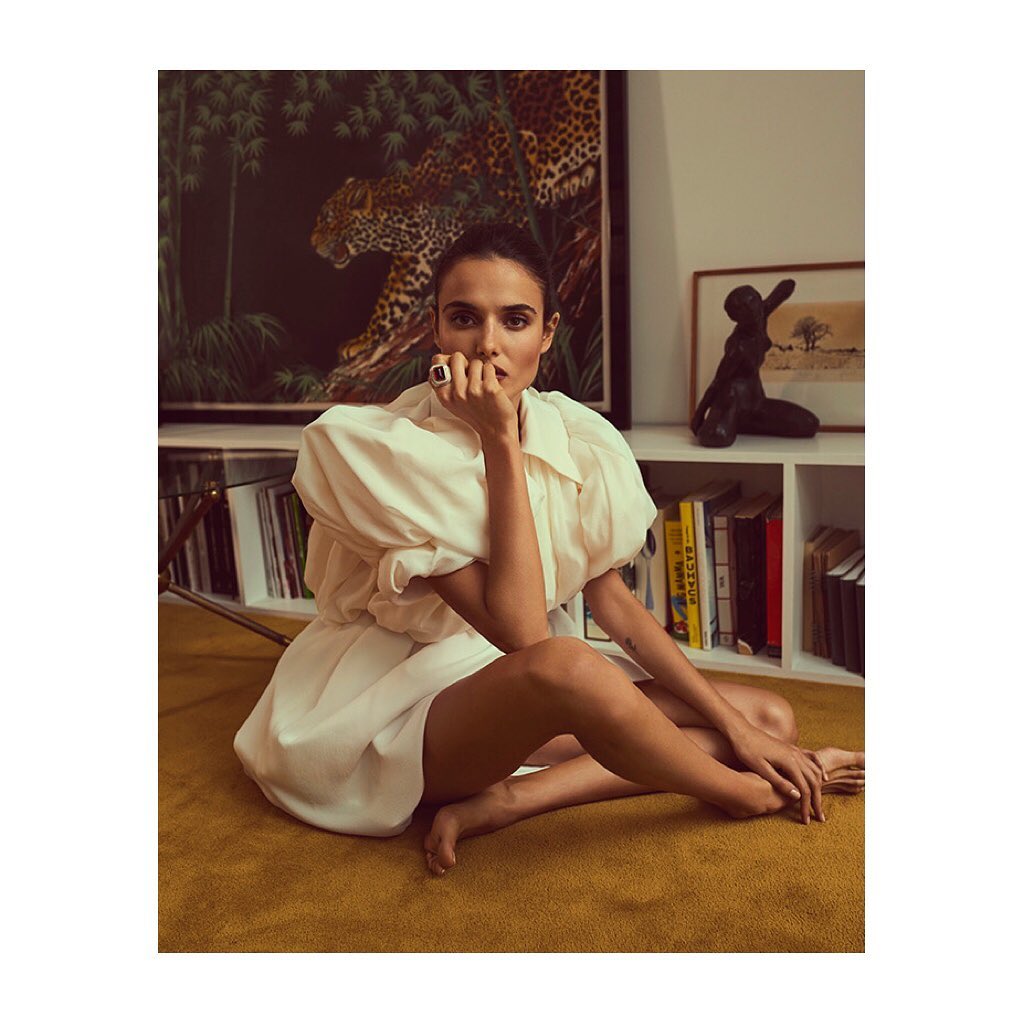 Brunette woman sits cross-legged on mustard-colored carpet wearing a large, puffed-sleeve cream dress, posing in an indoor setting with artwork and books.