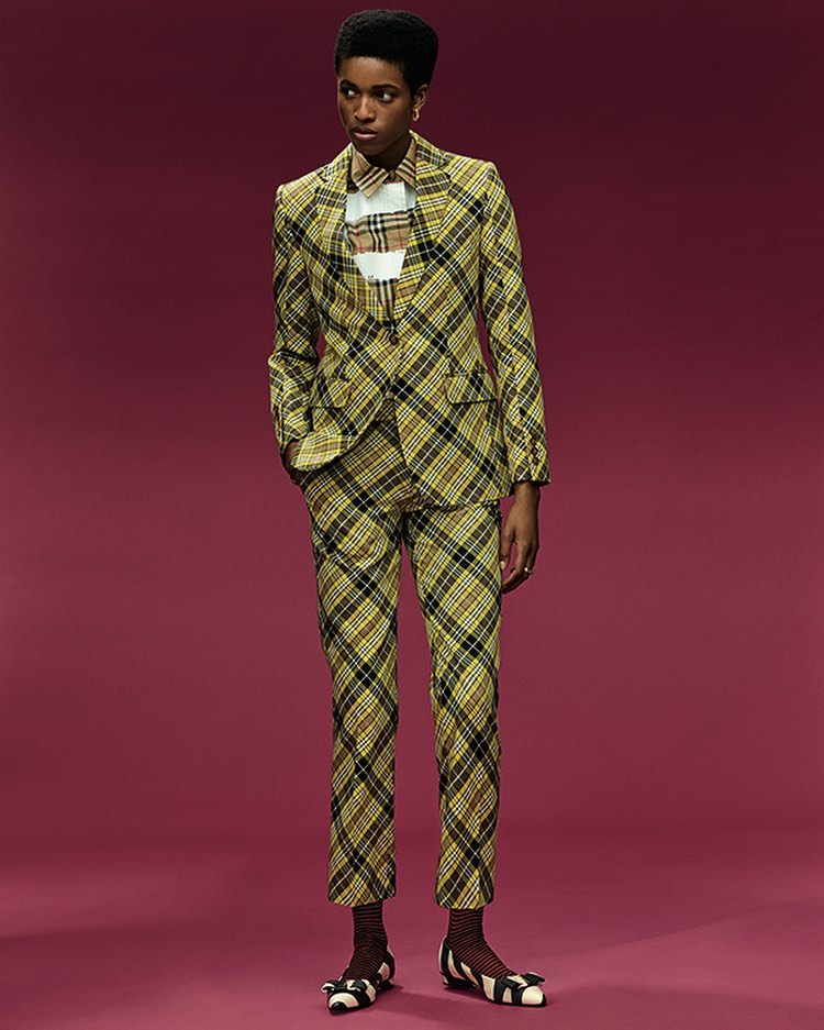 Woman poses in a plaid suit jacket and pants, accessorized with striped ballet flats against a solid purple-pink backdrop in studio lighting.
