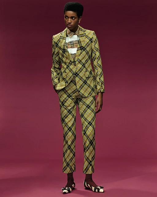 Woman poses in a plaid suit jacket and pants, accessorized with striped ballet flats against a solid purple-pink backdrop in studio lighting.