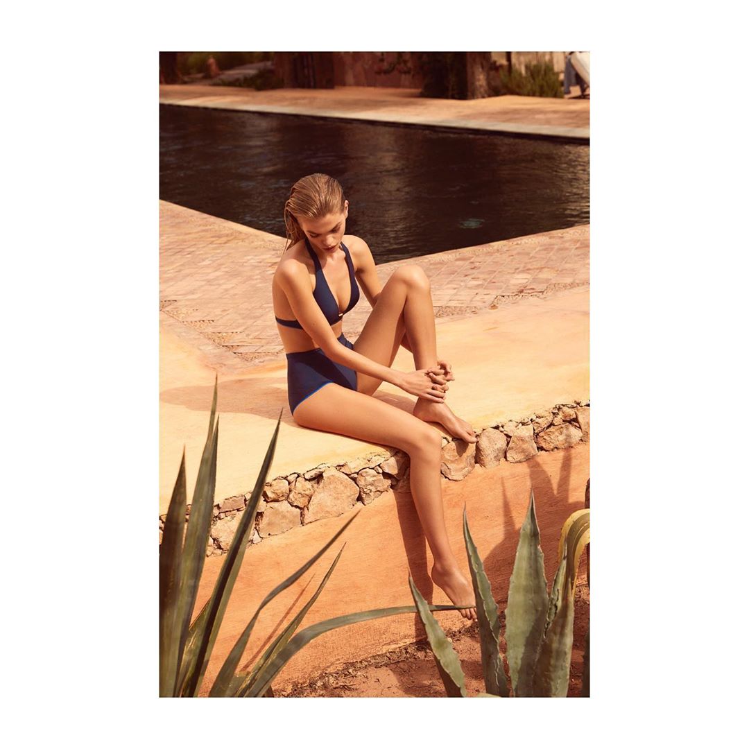A woman in a blue bikini sits on a stone ledge near a swimming pool, with desert plants in the foreground and a building in the background.