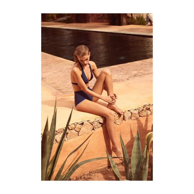 A woman in a blue bikini sits on a stone ledge near a swimming pool, with desert plants in the foreground and a building in the background.
