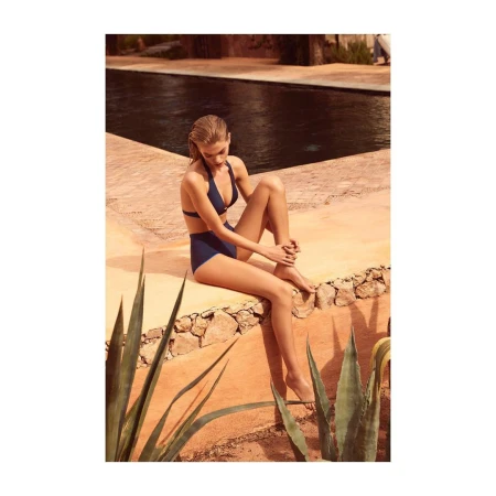 A woman in a blue bikini sits on a stone ledge near a swimming pool, with desert plants in the foreground and a building in the background.