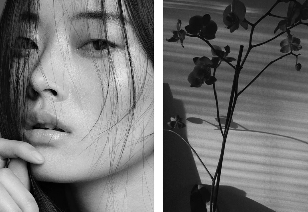 Close-up black and white portrait of an Asian woman with loose hair next to a flowering orchid branch against a textured wall.