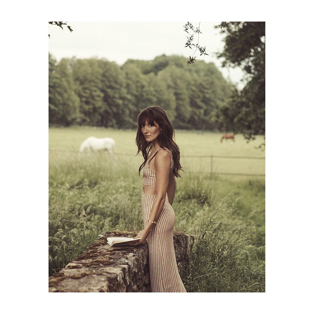 Adult female in striped dress stands by stone wall with a book in a green meadow with grazing horses on a cloudy day.