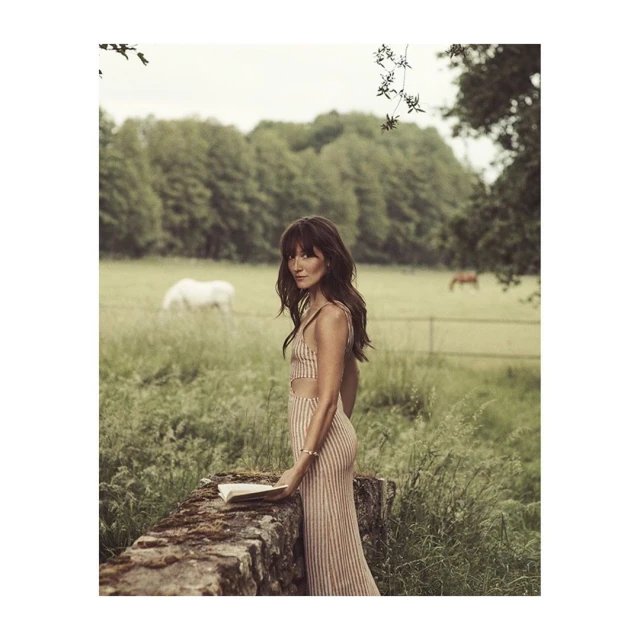 Adult female in striped dress stands by stone wall with a book in a green meadow with grazing horses on a cloudy day.
