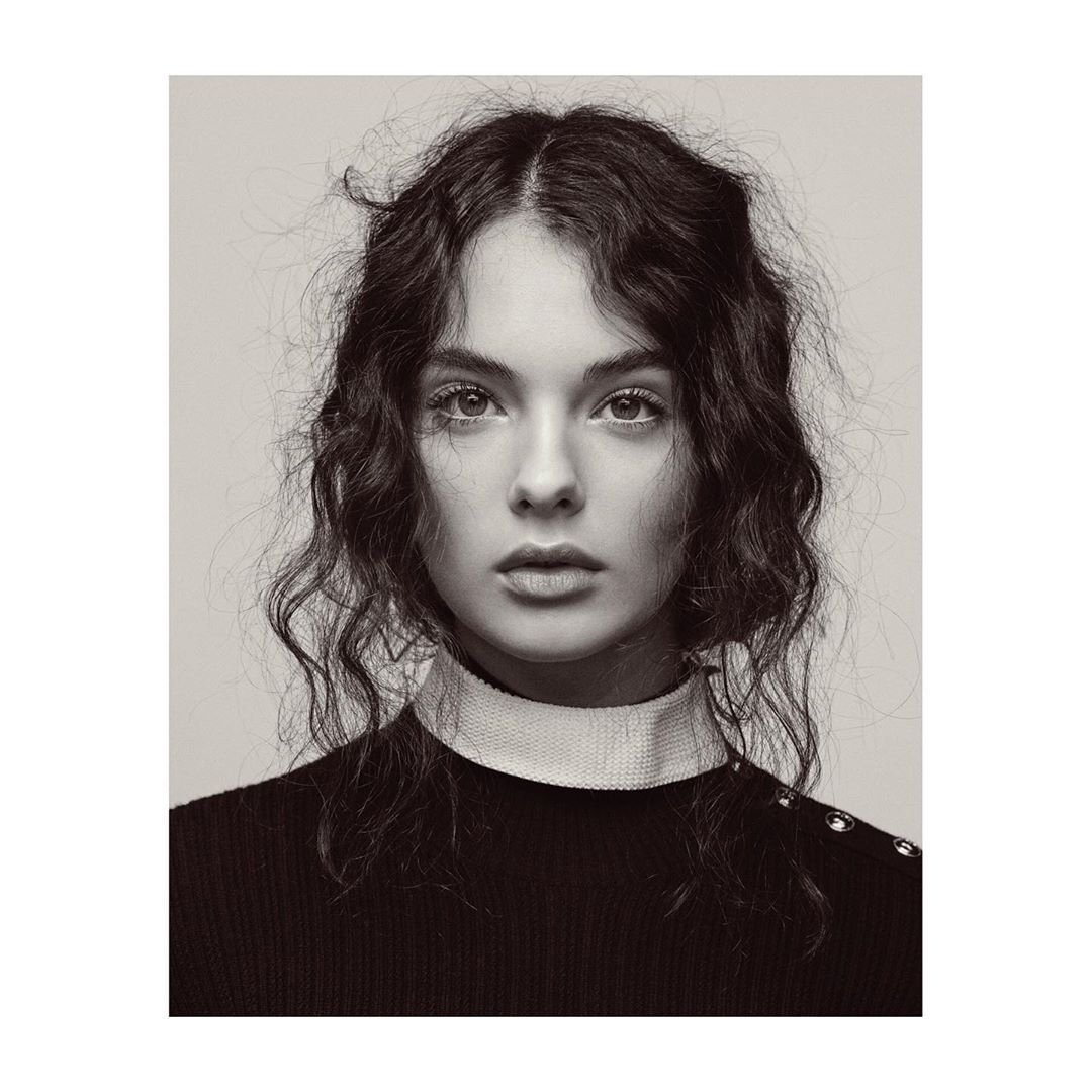 Portrait of a young woman with curly hair and a neutral expression, wearing a black sweater with white collar, in a black and white photo.