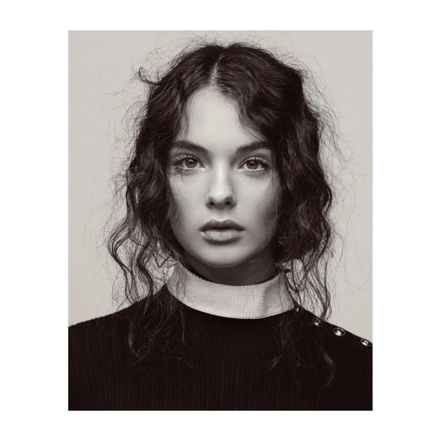 Portrait of a young woman with curly hair and a neutral expression, wearing a black sweater with white collar, in a black and white photo.