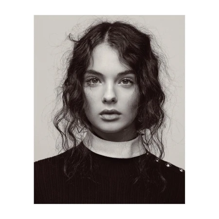 Portrait of a young woman with curly hair and a neutral expression, wearing a black sweater with white collar, in a black and white photo.