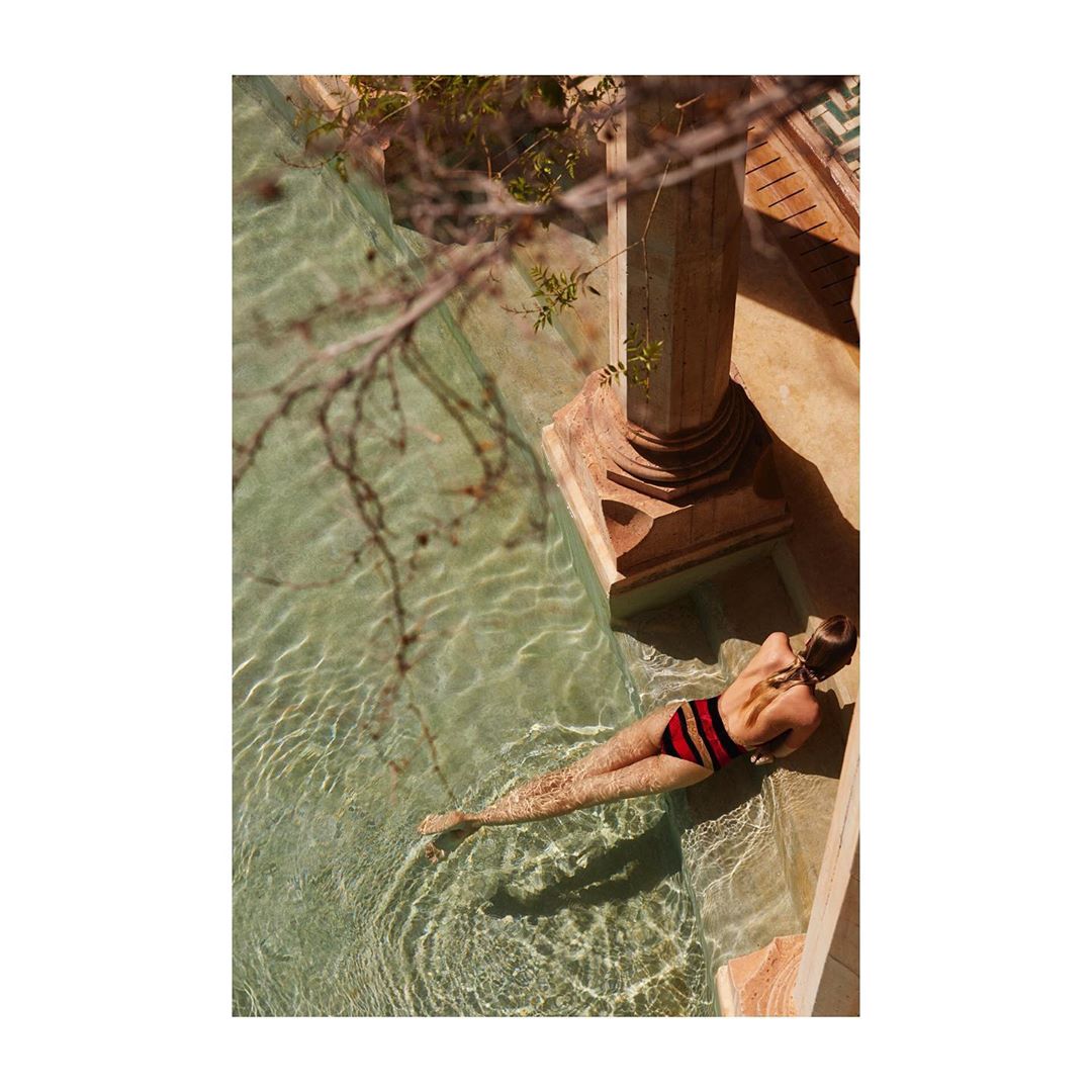 Woman in striped bathing suit lies in shallow rippled water of outdoor pool, sunlit and shaded, framed by stone and leafy branch.