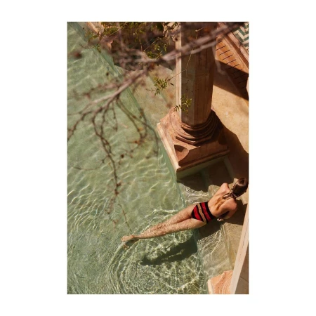 Woman in striped bathing suit lies in shallow rippled water of outdoor pool, sunlit and shaded, framed by stone and leafy branch.
