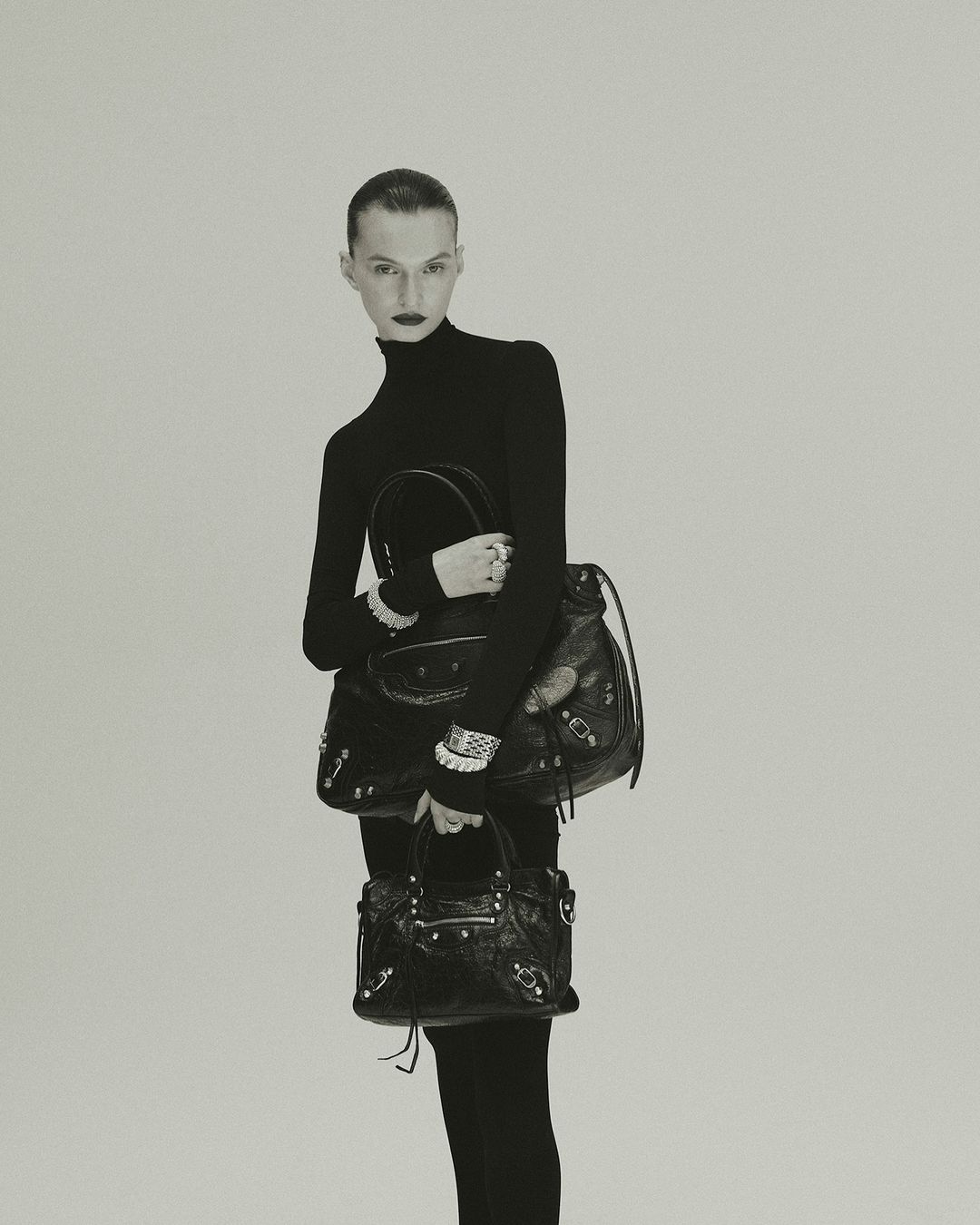 Model in black turtleneck holding two black leather handbags against a neutral studio background by Jonas Bresnan