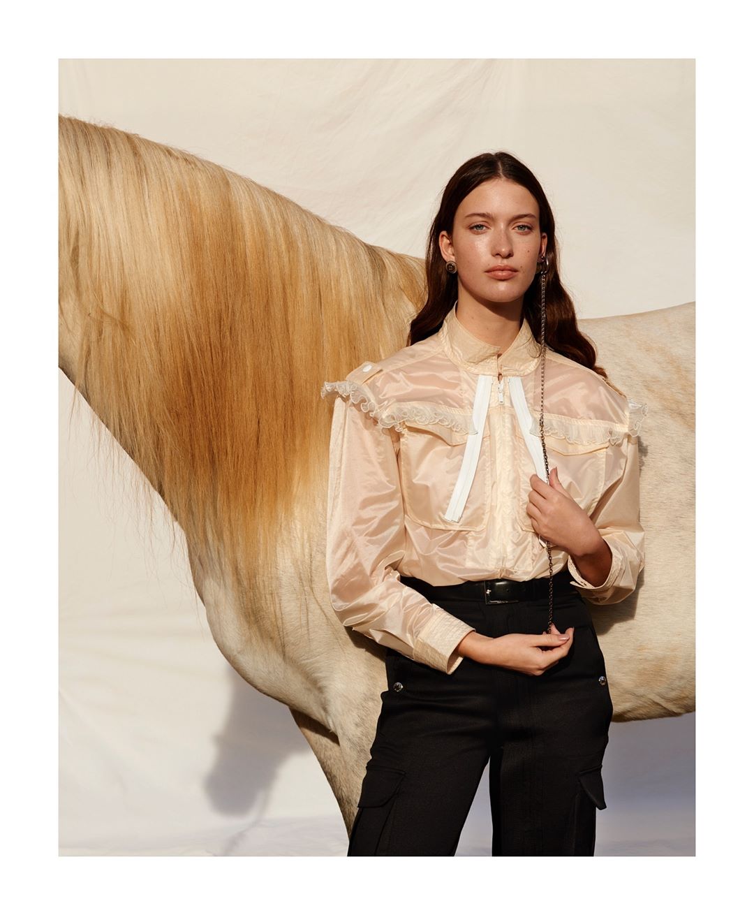 A woman in a sheer blouse poses with a light brown horse, looking directly at the camera in a studio environment.