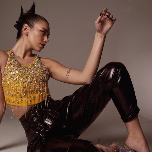 A woman with dark hair in a high-fashion pose wearing a yellow jeweled crop top and shiny black pants against a grey backdrop.