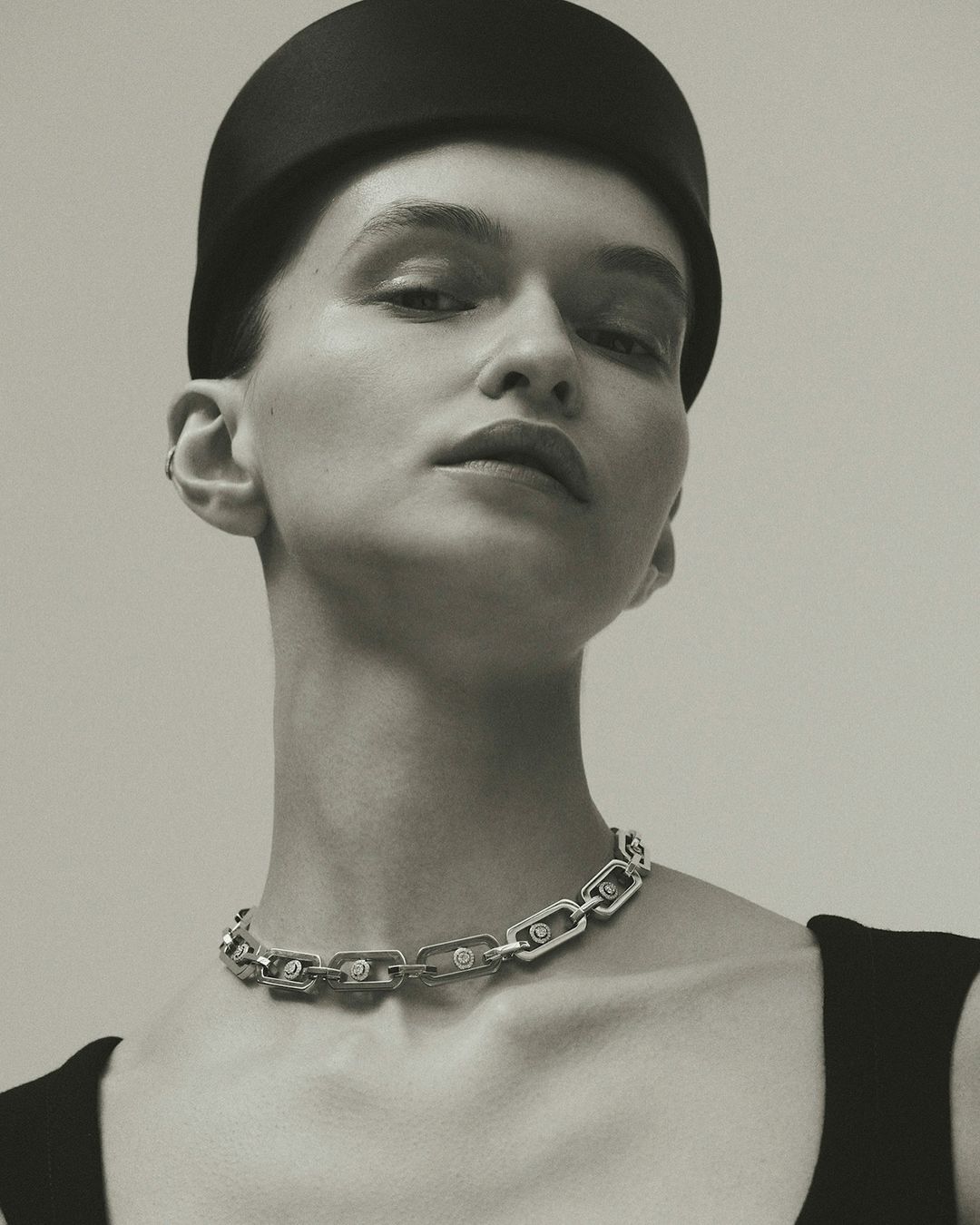 A sophisticated studio portrait shows a woman wearing a black hat and silver chain link necklace with diamond accents against a pale gray background.