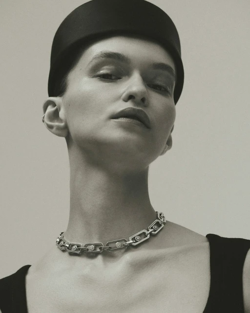A sophisticated studio portrait shows a woman wearing a black hat and silver chain link necklace with diamond accents against a pale gray background.