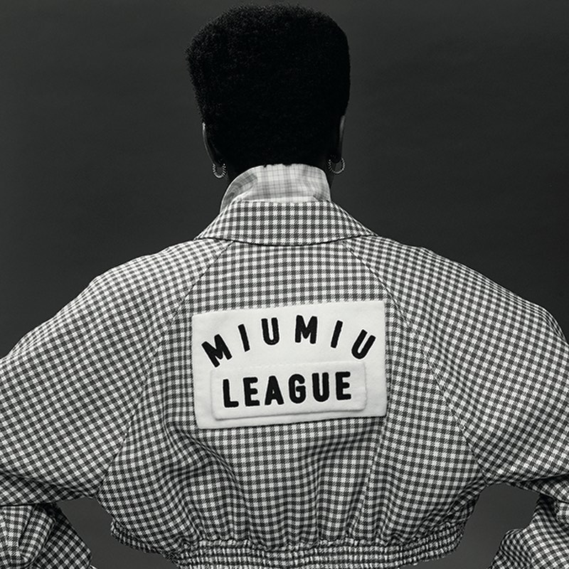 A woman with dark hair in a white and grey gingham jacket featuring a 'Miu Miu League' patch, captured in a back-facing studio shot.