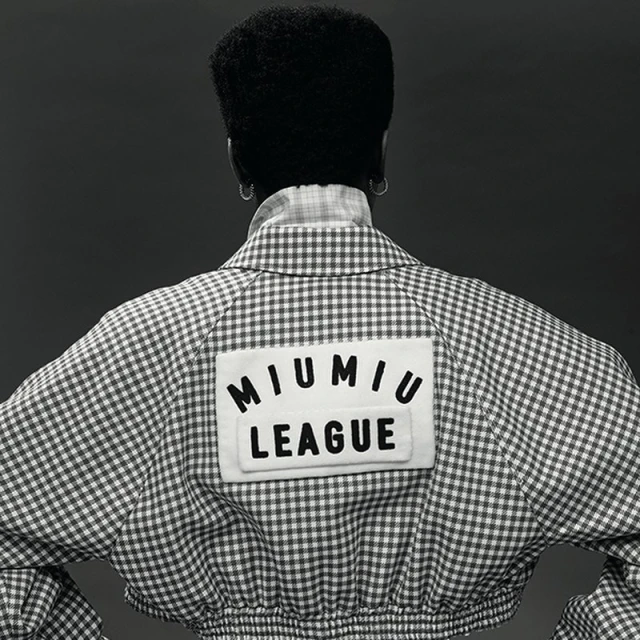 A woman with dark hair in a white and grey gingham jacket featuring a 'Miu Miu League' patch, captured in a back-facing studio shot.